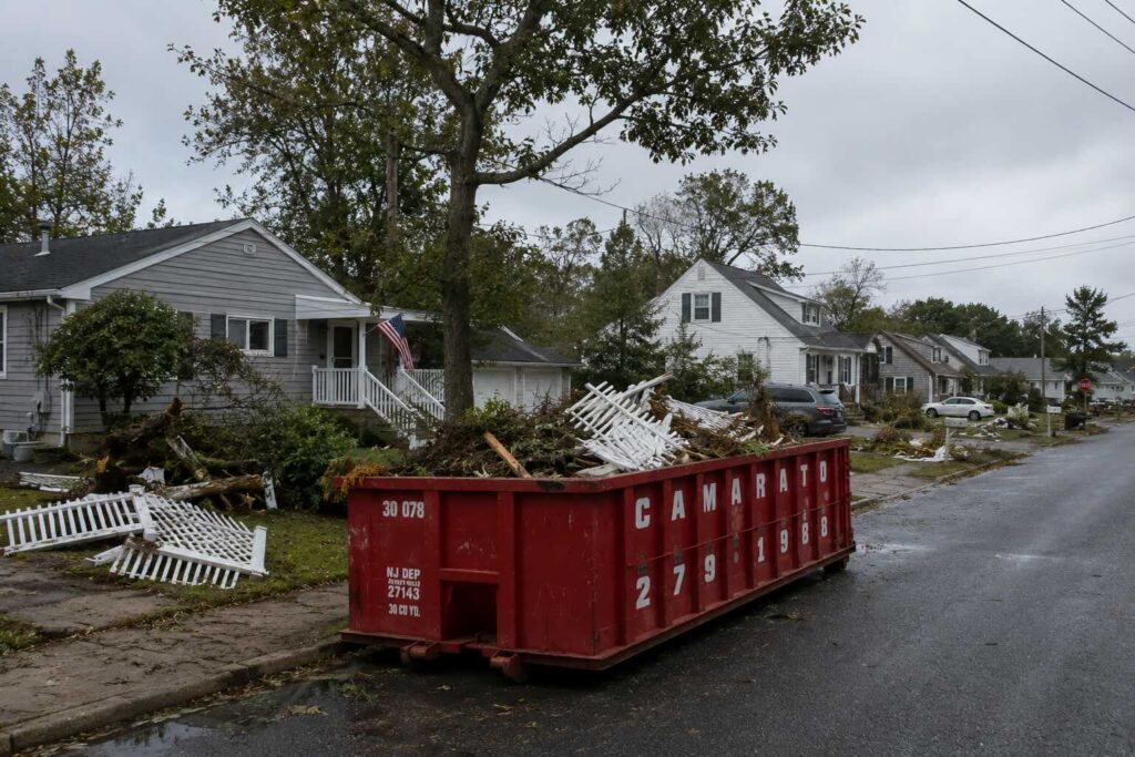 Roll-off dumpster filled with storm debris, broken fencing, and tree branches parked curbside in a coastal New Jersey residential neighborhood under an overcast post-storm sky.