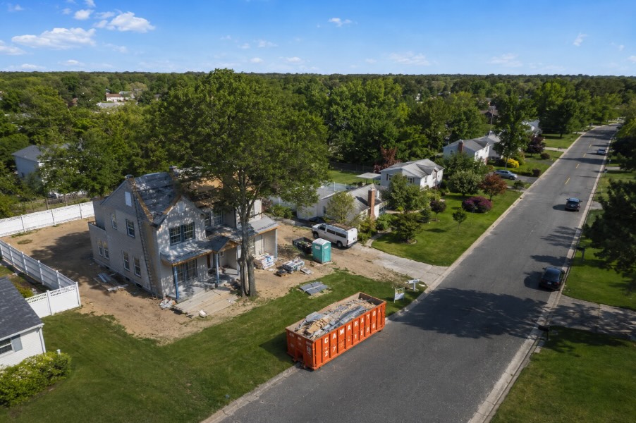 Aerial view of a suburban Ocean County, New Jersey neighborhood with tree-lined streets, single-family homes, and an orange roll-off dumpster placed curbside beside a home under renovation on a sunny morning.